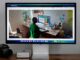 A computer monitor displays an image of a person working at a desk in a room filled with plants and various items. Below the monitor, a camera and a small computer device are placed on a wooden surface.