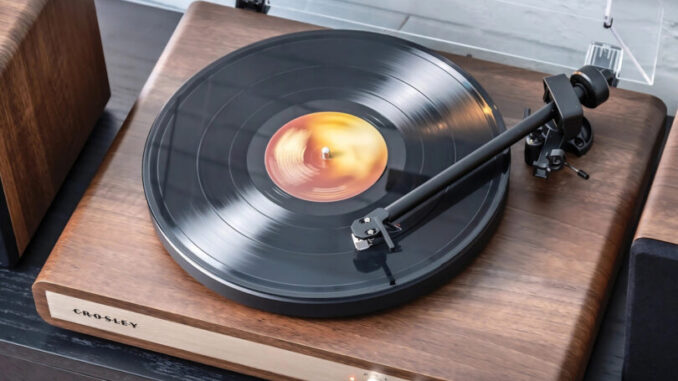 A close-up of a Crosley C65 wooden turntable playing a vinyl record next to a matching wooden speaker on a dark surface against a light brick wall.