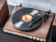 A close-up of a Crosley C65 wooden turntable playing a vinyl record next to a matching wooden speaker on a dark surface against a light brick wall.
