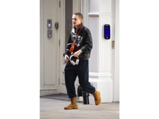 Timothée Chalamet walking on a street, wearing a black jacket, dark sweatpants, brown boots, and an orange shoulder bag, holding white headphones.