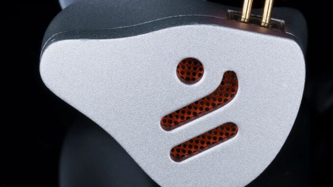 A Rising Hi-Fi Brand's New Audiophile Headphones Are More Affordable Close-up of a silver metallic device with a perforated orange grille and two gold connectors on top.