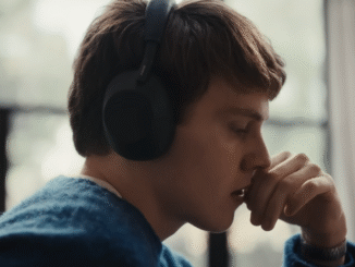 A young boy wearing a pair of blue headphones and blue jumper holding his hand up against his mouth
